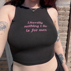 Pode incluir: Top preto com a frase bordada em rosa "Literally nothing I do is for men". O top é sem mangas, em estilo malha canelada.