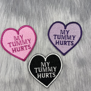 May include: Three heart-shaped iron-on patches with the text "My Tummy Hurts" in white embroidery. The patches are pink, purple, and black.