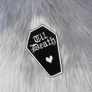 May include: A black coffin-shaped embroidered patch with a white border. The text "Til Death" is stitched in white using a gothic-style font, positioned above a small, simple white heart icon centred in the lower portion of the patch.