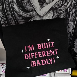 May include: Black t-shirt with pink embroidered text that reads "I'm built different (badly)" with white stars on either side of the text.