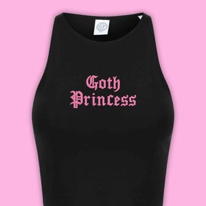 May include: A black sleeveless crop top featuring a high neckline and the text Goth Princess printed in a light pink gothic font across the chest. A white brand label with the letters SF is visible at the inside of the neckline.