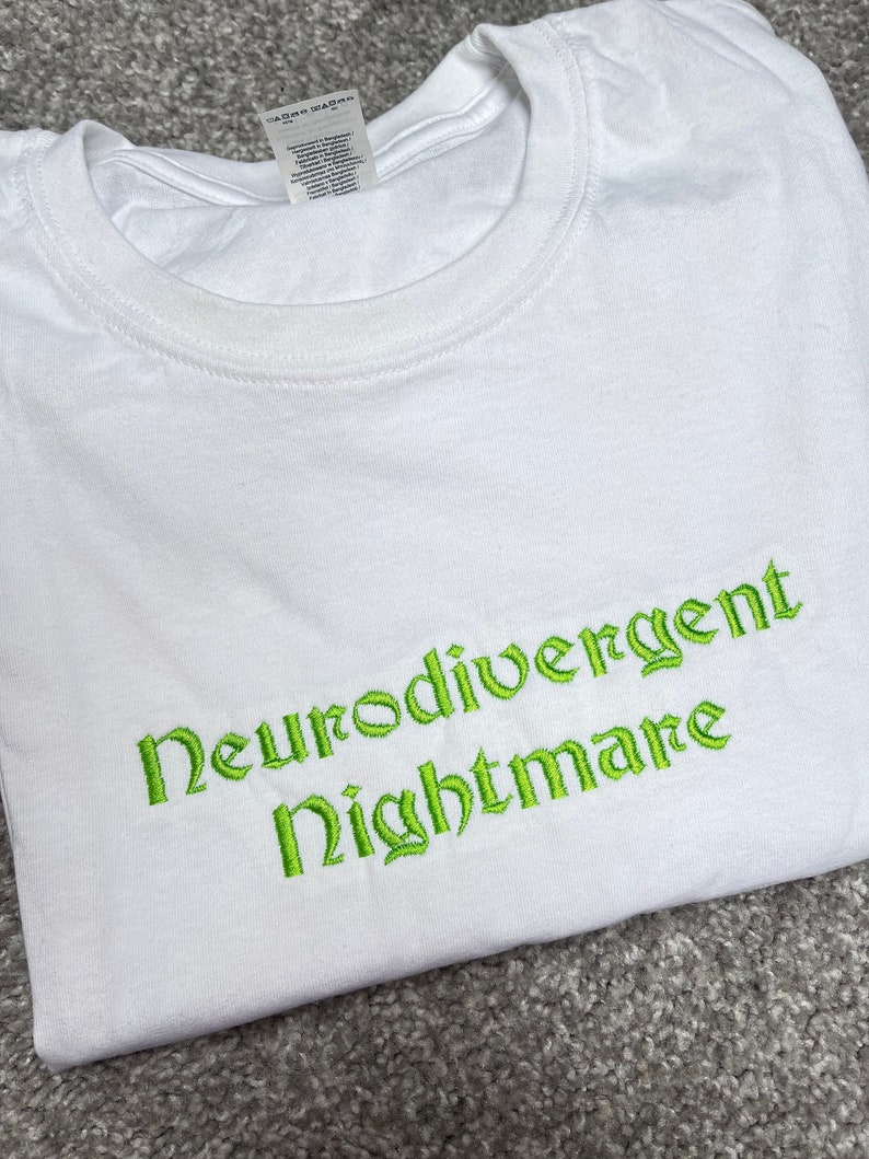 May include: White t-shirt with green embroidered text that reads "Neurodivergent Nightmare".