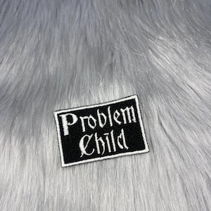 Op de afbeelding: Zwart rechthoekig embleem met de woorden "Problem Child" geborduurd in witte gotische letters. De patch heeft een witte rand en is geplaatst tegen een achtergrond van grijs imitatiebont.