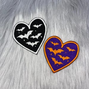 May include: Two heart-shaped embroidered patches. One patch is black with white embroidered bats and a white border. The second patch is purple with orange embroidered bats and an orange border. Both patches feature a repeating bat pattern.