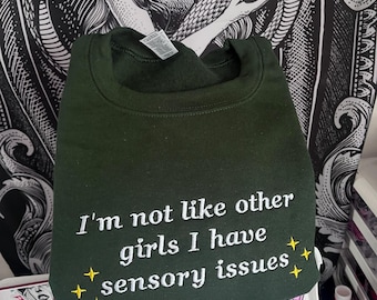 Könnte beinhalten: Ein dunkelgrüner Pullover mit dem Text "I'm not like other girls I have sensory issues" in weiß mit gelben Sternen gestickt.