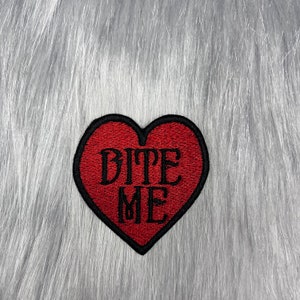 May include: Red felt heart-shaped patch with black embroidered text that reads "BITE ME".