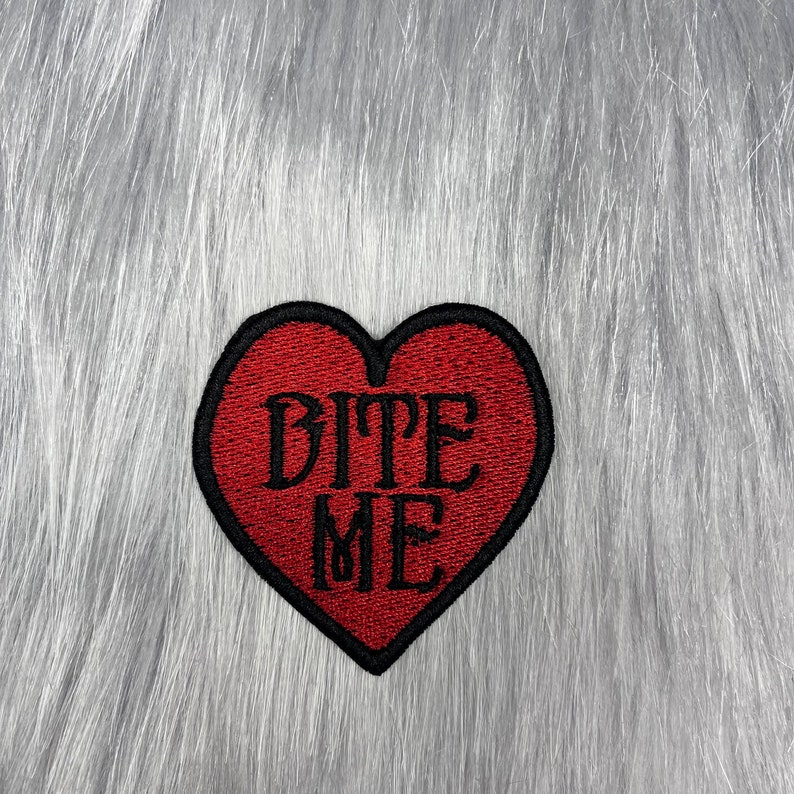 May include: Red felt heart-shaped patch with black embroidered text that reads "BITE ME".