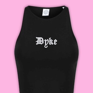 Könnte beinhalten: Ein schwarzes ärmelloses bauchfreies Oberteil mit hohem Ausschnitt und dem Wort "Dyke", das in weißer gotischer Schrift mittig auf der Brust gestickt ist. Das Kleidungsstück hat ein weißes Innenetikett am Kragen mit den Buchstaben "SF" in Schwarz.