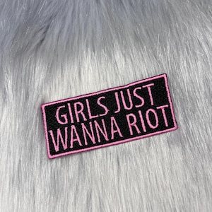 May include: A rectangular embroidered patch with a black background and a pink border. The text GIRLS JUST WANNA RIOT is stitched in pink capital letters across the centre of the patch, featuring a clean, bold, and sans-serif font style.