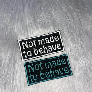 May include: Two rectangular embroidered patches with the text "Not made to behave." One patch has a black background with white text, and the other has a black background with teal text. The patches are displayed on a fluffy, light gray surface.