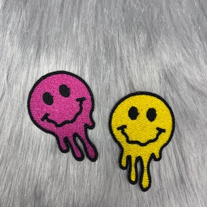 May include: Two embroidered iron-on patches in the shape of a smiley face with dripping edges. One patch is pink and the other is yellow.
