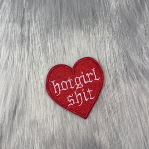 May include: A red heart-shaped embroidered patch with the words "hotgirl shit" in white gothic lettering. The patch is set against a backdrop of grey faux fur.