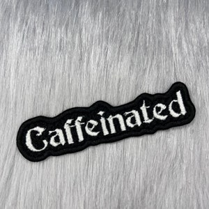 Puede incluir: Parche bordado en blanco y negro con la palabra "Caffeinated" en una fuente gótica.