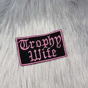 May include: A rectangular embroidered patch with a black background and a pink border. The text Trophy Wife is stitched in a pink gothic-style font across the centre of the patch. The patch has a textured surface and sharp, clean edges.