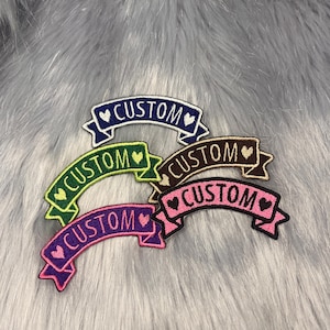 May include: Five embroidered patches in navy blue, green, brown, pink, and purple. Each patch has the word "CUSTOM" in white, with a heart on each side. The patches are on a grey, furry background.