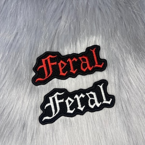 Może przedstawiać: Dwie haftowane naszywki ze słowem "Feral". Jedna naszywka ma czerwone litery na czarnym tle, druga ma białe litery na czarnym tle. Naszywki znajdują się na szarej, futrzanej powierzchni.