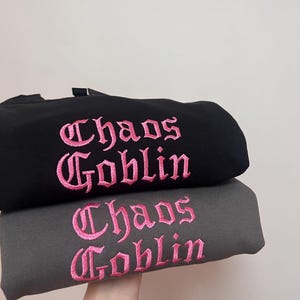 May include: Two sweatshirts stacked on top of each other. The top sweatshirt is black with pink embroidered text that says "Chaos Goblin". The bottom sweatshirt is gray with pink embroidered text that says "Chaos Goblin".