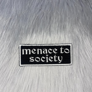 Può includere: Una toppa rettangolare nera con testo ricamato bianco che recita "menace to society". La toppa ha un bordo bianco ed è ambientata su uno sfondo di finta pelliccia grigia.