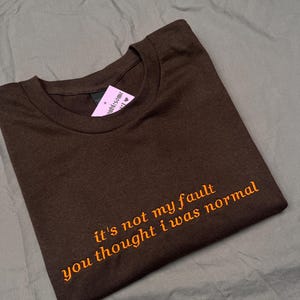 Op de afbeelding: Een gevouwen bruin t-shirt met de geborduurde tekst "it's not my fault you thought I was normal" in oranje. Het shirt heeft een ronde hals en een label.