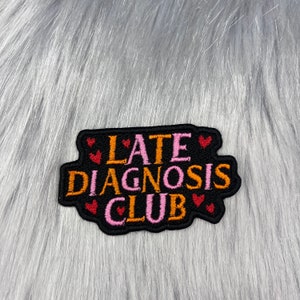 May include: Black embroidered patch with the text "Late Diagnosis Club" in orange and pink lettering. The patch is surrounded by small pink and red hearts.