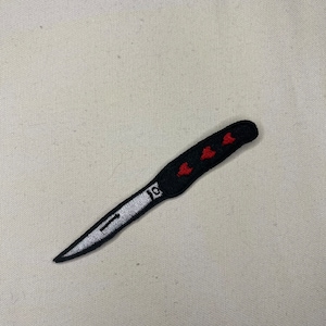 May include: Black and white embroidered patch of a knife with a red heart design on the handle.