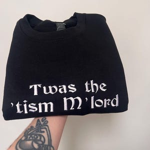 May include: Black t-shirt with white embroidered text that reads "Twas the 'tism M'lord"