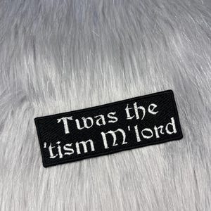 May include: Black rectangular patch with white embroidered text that reads "Twas the 'tism M'lord". The patch is set against a backdrop of gray faux fur.