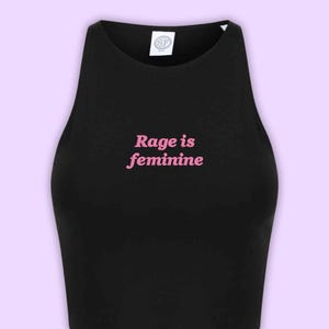 Op de afbeelding: Zwarte crop top met de roze tekst "Rage is feminine" in het midden. De top heeft een ronde hals en is mouwloos. Een klein wit label is zichtbaar bij de halslijn.