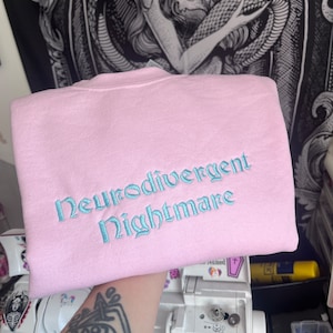 May include: A pink sweatshirt with the text "Neurodivergent Nightmare" embroidered in light blue.
