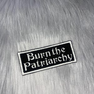 Peut inclure: Un écusson rectangulaire noir avec le texte brodé en blanc « Burn the Patriarchy ». L'écusson a une bordure blanche et est posé sur un fond de fausse fourrure grise.