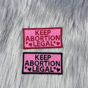 May include: Two pink fabric patches with black stitching. The text on the patches reads "Keep Abortion Legal" with a small heart on either side of the word "Legal".