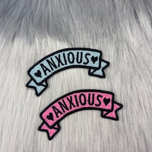 May include: Two embroidered iron-on patches with the word "ANXIOUS" in black lettering on a banner with a heart at each end. One patch is light blue and the other is pink.
