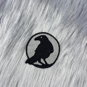 Puede incluir: Un parche circular gris con la silueta de un cuervo negro bordada. El parche es perfecto para añadir un toque de estilo gótico a la ropa, bolsos u otros accesorios.