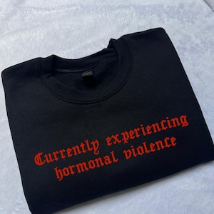 May include: Black sweatshirt with the phrase "Currently experiencing hormonal violence" in red gothic font. The sweatshirt is laid flat on a white, textured surface.