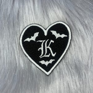 May include: A heart-shaped embroidered patch with a black background and a white border. The centre features a large white letter K in a gothic font, surrounded by three small white embroidered bats positioned above and below the letter.