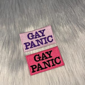 Könnte beinhalten: Zwei gestickte Patches mit dem Text "Gay Panic" in verschiedenen Farben. Der obere Patch ist rosa mit lilafarbenem Text. Der untere Patch ist rot mit schwarzem Text.