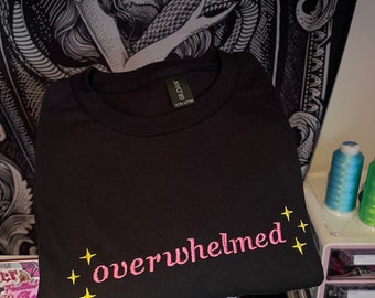 May include: Black t-shirt with the word "overwhelmed" embroidered in pink, accompanied by yellow star accents. The shirt is folded and appears to be made of a soft material. The brand tag is visible at the neckline.