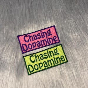 Puede incluir: Dos parches bordados con el texto "Chasing Dopamine" en diferentes colores. El parche superior es rosa con texto morado y el parche inferior es amarillo con texto verde.