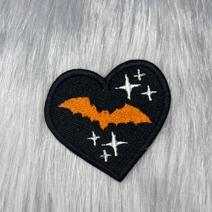 Puede incluir: Parche de fieltro negro en forma de corazón con un murciélago naranja y estrellas blancas bordadas.