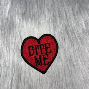 Puede incluir: Parche de fieltro rojo en forma de corazón con texto bordado en negro que dice "Bite Me".