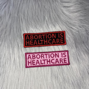 May include: Two rectangular embroidered patches featuring the text ABORTION IS HEALTHCARE. The top patch has black lettering on a red background with a black border. The bottom patch has pink lettering on a pink background with a pink border.