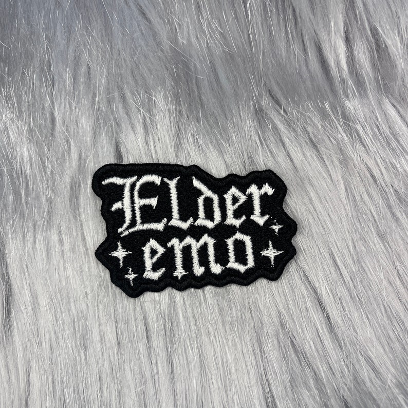 May include: Black embroidered patch with white lettering that reads "Elder emo" with two white stars on either side of the text.