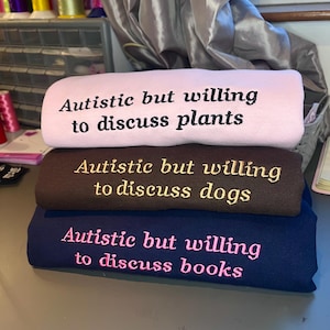 May include: Three folded sweatshirts, stacked. The top one is light pink with the embroidered text "Autistic but willing to discuss plants." The middle one is brown with "Autistic but willing to discuss dogs." The bottom one is navy blue with "Autistic but willing to discuss books."