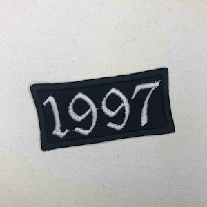Pode incluir: Etiqueta bordada preta com costuras brancas, apresentando o ano "1997" em uma fonte estilizada.