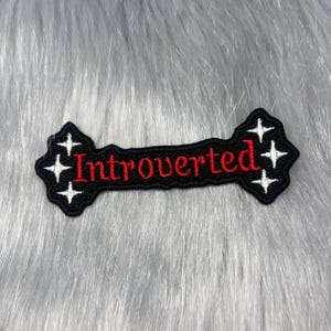 May include: A black embroidered patch with a scalloped edge featuring the word Introverted stitched in red thread. The design includes white four-pointed star accents on both the left and right sides of the text, creating a decorative border.