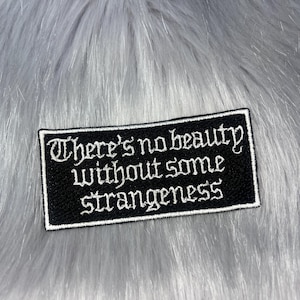 May include: A rectangular black embroidered patch with a white border and white text that reads, There's no beauty without some strangeness. The patch features a gothic-style font and is displayed against a soft, light grey faux fur background.