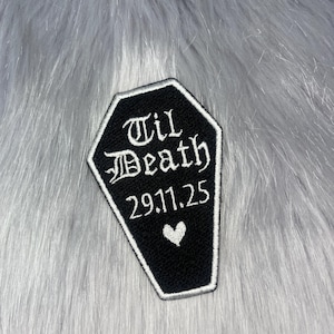 Puede incluir: Un parche bordado con forma de ataúd con fondo negro y borde blanco. El parche presenta el texto bordado en blanco "Til Death" en una fuente gótica, seguido de la fecha "29.11.25" y un pequeño icono de corazón blanco debajo.