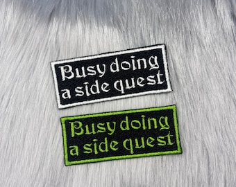 May include: Two black fabric patches with white and green embroidery. The text on the patches reads "Busy doing a side quest".