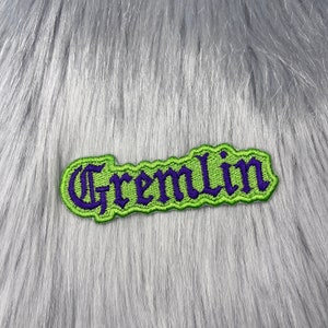 Può includere: Toppa ricamata verde e viola con la parola "Gremlin" in un font gotico.