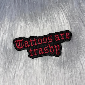 May include: A black embroidered patch with a jagged, irregular border. The text "Tattoos are trashy" is stitched in a bold, red, gothic-style font across the centre of the patch, creating a high-contrast design with the black background.